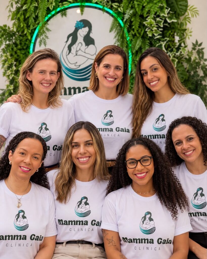 Equipe multidisciplinar da Clínica Mamma Gaia, em São Paulo, reunida em frente ao painel verde com o logo da clínica